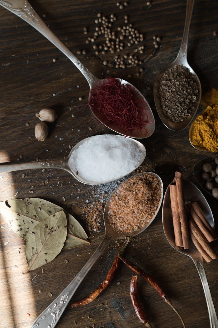 Top-down view of various spices and herbs in spoons on a rustic wooden table.